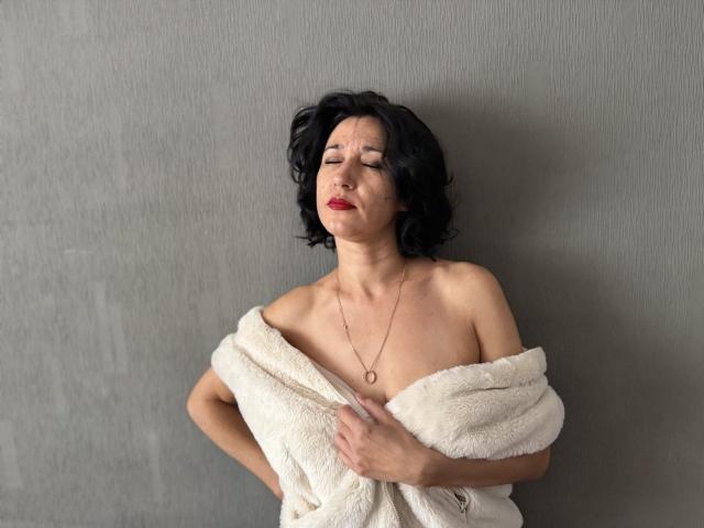 CameronEnns with black curly hair, eyes closed, red lipstick, white fluffy towel covering her tits, holding it with one hand, necklace, gray wall, slightly leaning back, looking relaxed.