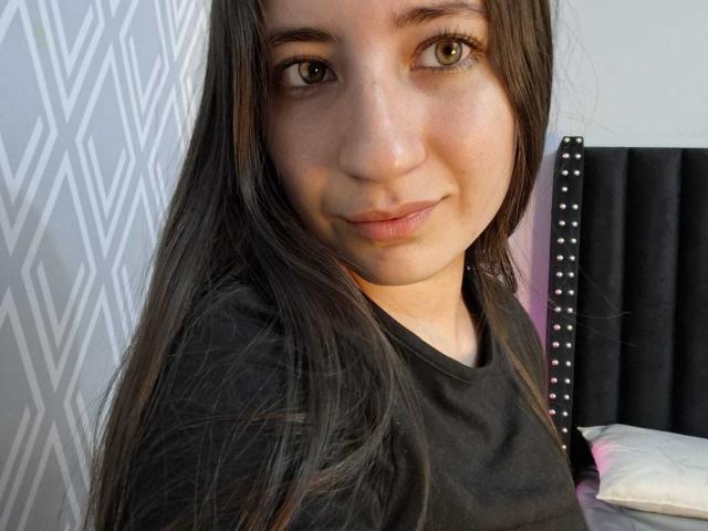 HottieDayCare hat langes, braunes Haar, grüne Augen und ein süßes Lächeln. Sie trägt ein schwarzes T-Shirt, und ihre kleinen Brüste sind leicht sichtbar. Das Bett hat ein schwarzes Kopfteil..