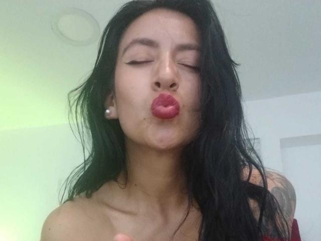 ZarayFeet with closed eyes, pouty red lips, and long black hair, wearing pearl earrings, making a kissy face. Her bare shoulders and tattoo are visible.