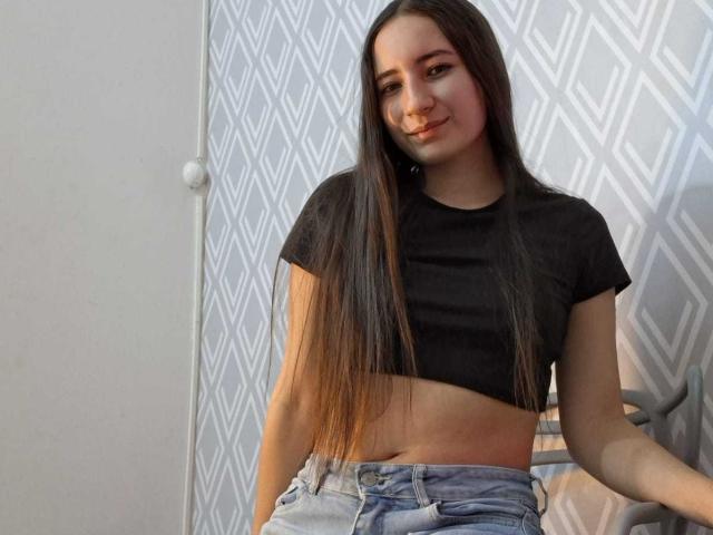 Una ragazza attraente con lunghi capelli castani, un top corto nero e jeans blu, seduta e sorridente, che mostra un po' l'addome e ha piccoli seni..