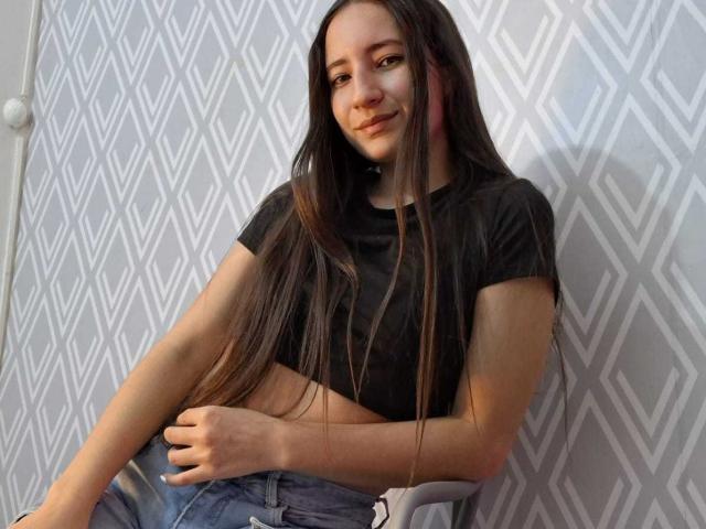 Creche com uma mulher atraente, com cabelo castanho longo, top cropped preto e jeans azuis, sentada em uma cadeira, sorrindo, mostrando suas nádegas planas, com a mão na barriga, em um quarto com padrão cinza..