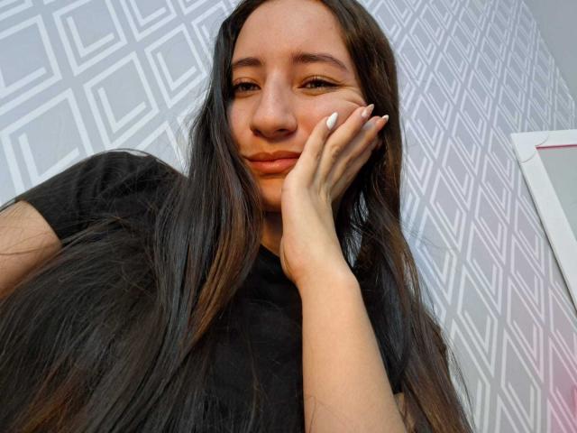 Una ragazza di HottieDayCare, con lunghi capelli neri, una maglietta nera e smalto bianco sulle unghie, appoggia il mento sulla mano e sorride leggermente. Ha il seno piccolo ed è carina..