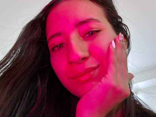 Creche Hottie com pele levemente rosada, cabelo longo e escuro, unhas brancas, com um sorriso discreto, com a mão apoiada no rosto, olhos castanhos, maquiagem suave, luz suave, selfie casual..