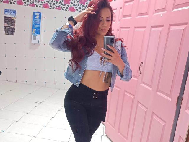 Emily Durand mit roten Haaren, die eine blaue Jeansjacke, ein weißes Crop-Top und schwarze, hochgeschnittene Hosen trägt, macht ein Selfie vor einem Spiegel und zeigt dabei ihr Gesäß und ihre schlanke Taille..