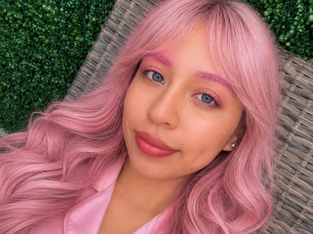 ZoeBell con cabello rosa, cejas rosas, párpados rosas y labios rosas, sonriendo a la cámara, vistiendo una camisa rosa, una silla de mimbre en el fondo, linda y sexy..
