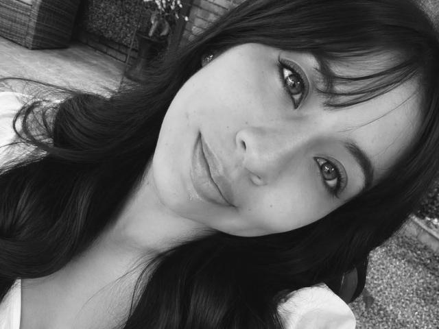 ZoeBell tiene el pelo largo y oscuro, piel clara y una ligera sonrisa con labios carnosos. Tiene ojos oscuros, ligeramente levantados, y lleva una blusa blanca. Foto en blanco y negro..