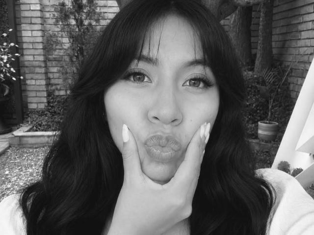 Black and white selfie of ZoeBell with long dark hair, pouty lips, and manicured nails, touching her face with her hand, showing off her ass-shaped lips.