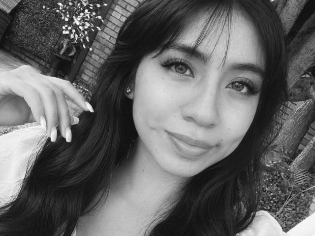 O rosto de ZoeBell, uma selfie em preto e branco, sorrindo com cabelos longos e escuros, cílios, brincos, camisa branca, unhas bem cuidadas, franja levemente bagunçada, pele macia, fofa, feminina, atraente..
