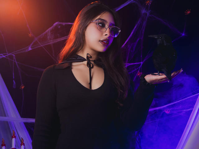 MelanyBlom with glasses, black top, and cleavage, holding a black crow, her lips slightly parted, dark wavy hair, purple and orange lighting, looking sexy and mysterious.