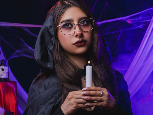 MelanyBlom with glasses, holding a lit candle, wearing a black hooded jacket, has long brown hair, and is looking directly at the camera with dark red lipstick.