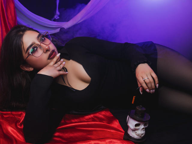 MelanyBlom with glasses, dark hair, red lipstick, black cleavage dress, black tights, lying on red silk, holding drink with straw, skull cup, purple blue light, looking at camera.