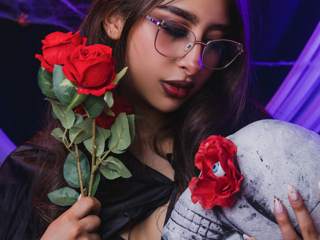 MelanyBlom with red roses, holding one in her hand, wearing glasses, black top, and gray patterned pants, looking down with pouty lips, purple lighting in background.