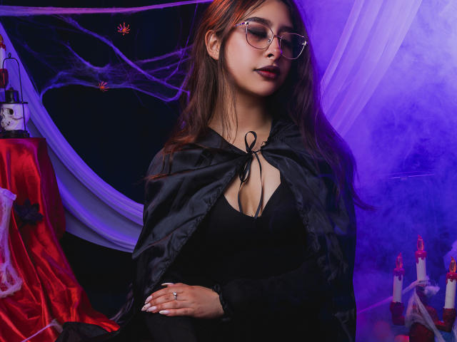 MelanyBlom with glasses, black cleavage dress, black cape, red lipstick, long brown hair, ring on finger, purple and blue lighting, red and black fabric, candles in background.