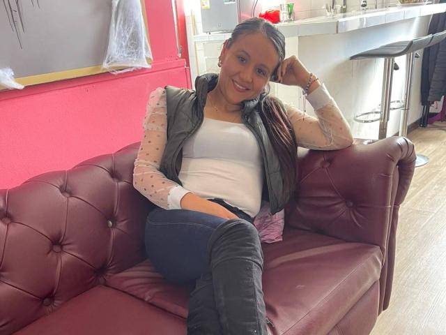 Liliana Tavera, con senos pequeños, vistiendo una blusa transparente con lunares, un chaleco negro, una camiseta blanca y jeans oscuros, sentada en un sofá de cuero púrpura, sonriendo a la cámara..