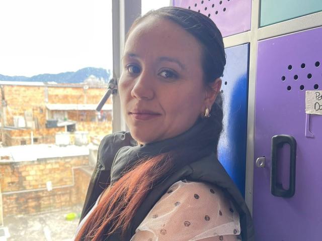 Liliana Tavera, com cabelo ruivo trançado, vestindo um colete verde sobre uma blusa branca transparente com bolinhas pretas, sorrindo para a câmera. Seus seios estão parcialmente visíveis..
