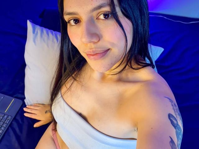 SophiaMarx, topless with white strap covering her tits, sitting on bed, laptop on left, blue and purple lights, black hair, tattoo on right arm, slight smile, fair skin.