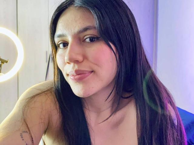 SophiaMarx with long black hair, light skin, small nose, slight smile, pink lips, tattoo on right arm, ring light on left, topless, purple and blue light reflections.