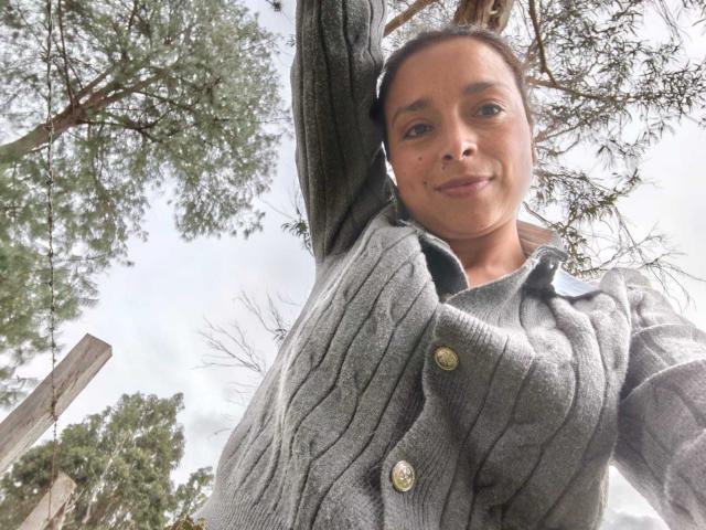 StangelarRivera with raised arm, wearing a gray cable-knit sweater, smiling up at the camera. Her tits are small, and she has medium brown skin and dark brown hair.