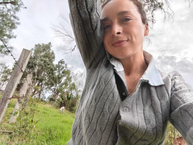 StangelarRivera with medium tits, wearing a gray ribbed sweater, arm raised, smiling, outdoors, grass, trees, cloudy sky, brown hair, light skin, slight nose, casual selfie.