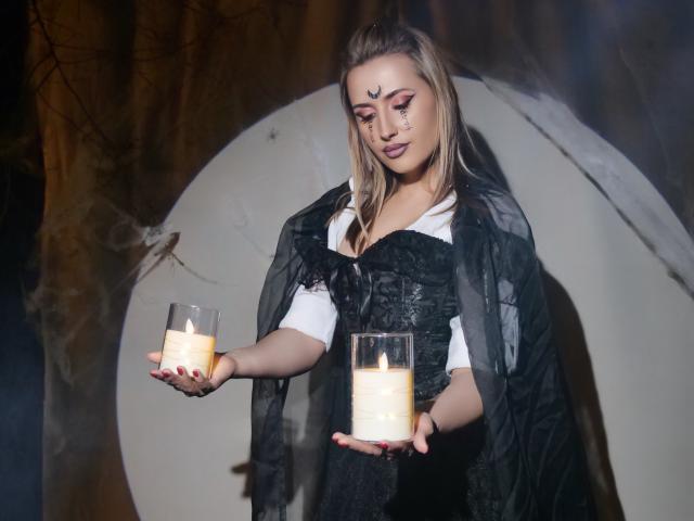 PassionGlow with blonde hair, black corset, and white shirt, holding two lit candles, has makeup with a black forehead mark, wearing a black sheer cape, looking down.