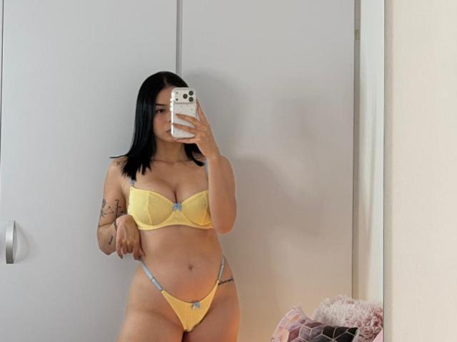 IsabellaJames with black hair, yellow bra, and yellow panties, holding a phone, showing her tits and pussy, tattoos on her left arm, mirror selfie, white wall behind.