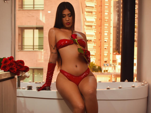NikaFoxxyHot with long black hair, red latex bra and panties, red gloves, holding rose, sitting in white tub, city skyline in background, rose bouquet on left.