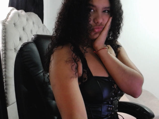AriaVega with curly black hair, wearing a black leather corset, sits in a black leather chair, hand on cheek, looking at the camera with a slight smile.