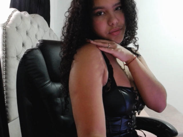 AriaVega with curly black hair, wearing a black leather corset, sits in a black leather chair, hand on neck, smiling slightly, showing her tits and a confident look.