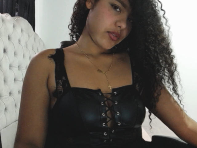 AriaVega, com cabelos pretos cacheados, vestindo um corpete preto com cadarços, exibindo seus seios e um colar com uma cruz dourada, sentada em uma cadeira branca com estofamento..