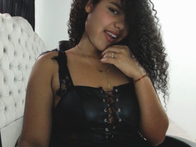 AriaVega with curly hair, smiling, wearing a black lace-up corset, showing her tits, gold necklace, and ring, sitting on a white tufted bed.