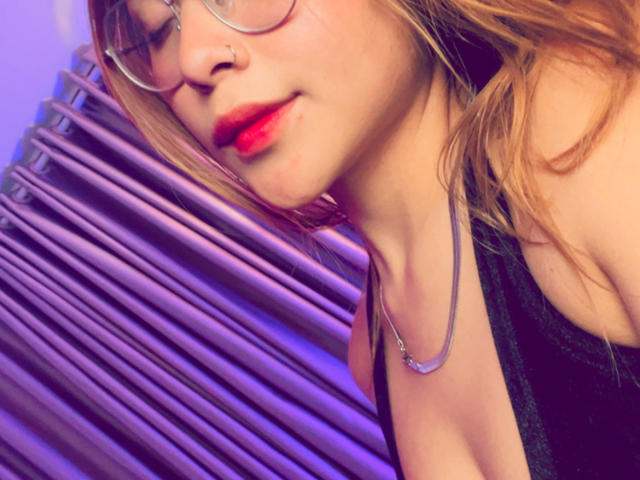 HeraJuno mit rotem Lippenstift, Brille und Nasenring, trägt ein schwarzes Oberteil, das ein wenig ihrer Brust zeigt, vor lila Vorhängen. Ihr blondes Haar ist leicht zerzaust..