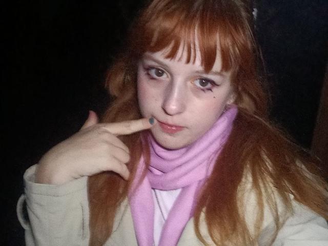 LouieOrner has pale skin, red hair, and is wearing a pink scarf with a white shirt and beige jacket. She's pointing at her slightly parted, glossy lips with her finger.