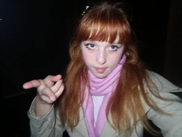 LouieOrner with red hair, pink scarf, and white top, making a "rock on" gesture, sticking out tongue, and showing off small, cute tits. Dark background.