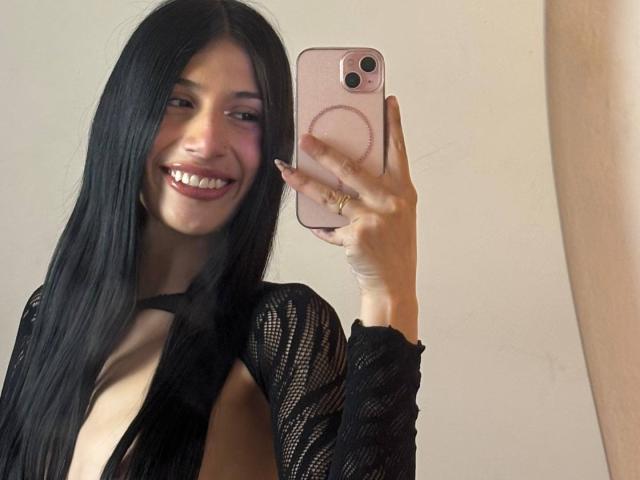 SophieBeaumont with long black hair, smiling, wearing a sheer black top, exposing her tits, holding a pink phone, taking a mirror selfie.