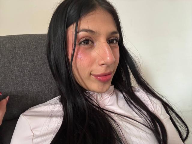 SophieBeaumont with long black hair, pinkish-red cheeks, slight smile, wearing white shirt, sitting in grey chair, pink lipstick, brown eyes, subtle freckles, natural light, soft skin.
