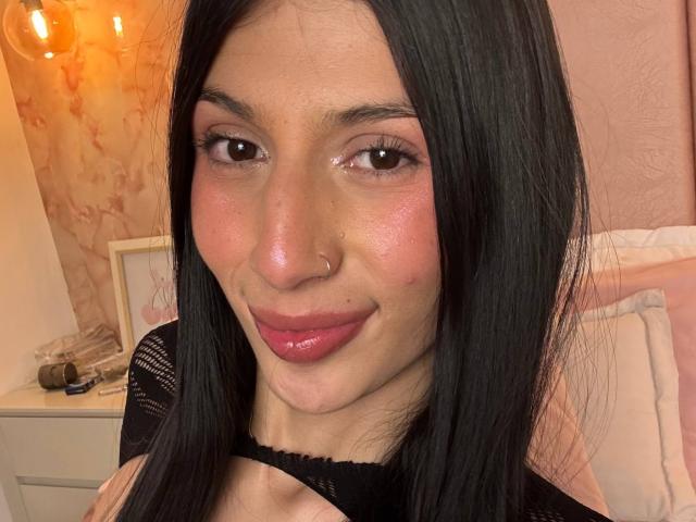 SophieBeaumont has shiny pink lips, black straight hair, brown eyes, and a cute nose ring. She's wearing a black lace top, looking cute with a subtle smile.