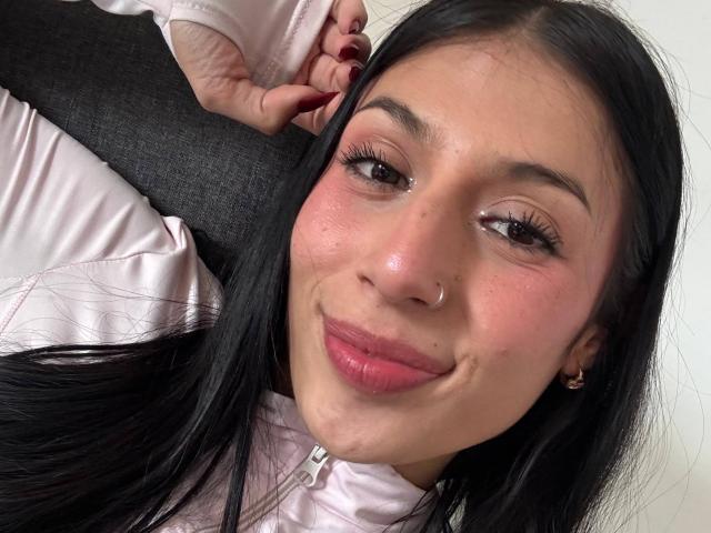 SophieBeaumont has black hair, pink lips, and a nose ring. She's smiling with her hand in her hair, wearing a pink jacket. Her pink cheeks are slightly shiny.