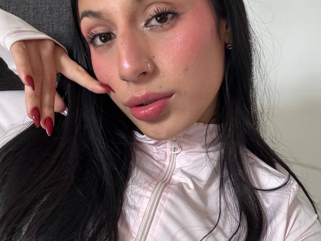 SophieBeaumont has shiny pink cheeks, long black hair, red nails, and a nose ring. She's wearing a white zip-up jacket, looking at the camera with a slight pout.