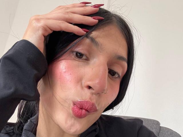 SophieBeaumont with puffy pink cheeks, black hair, red nails, nose ring, and puckered lips, hand in hair, wearing black long-sleeve shirt, looking into camera.