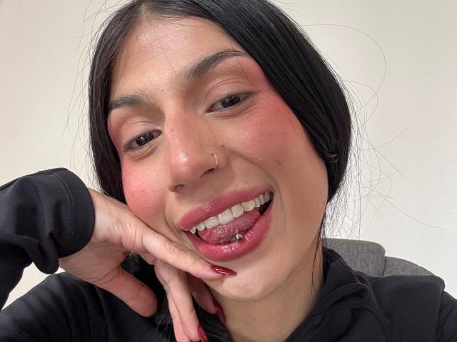 SophieBeaumont has a nose ring, black hair, red lipstick, and a tongue piercing with her hand on her cheek, smiling with red nails, wearing a black shirt.