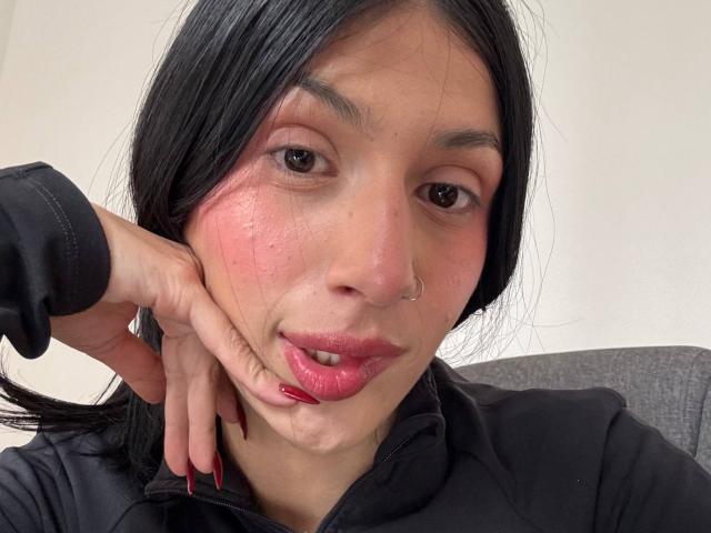 SophieBeaumont with black hair, nose ring, pink cheek, and red lipstick, pouting with hand on cheek, wearing black jacket, pink nail polish, and sitting in gray chair.