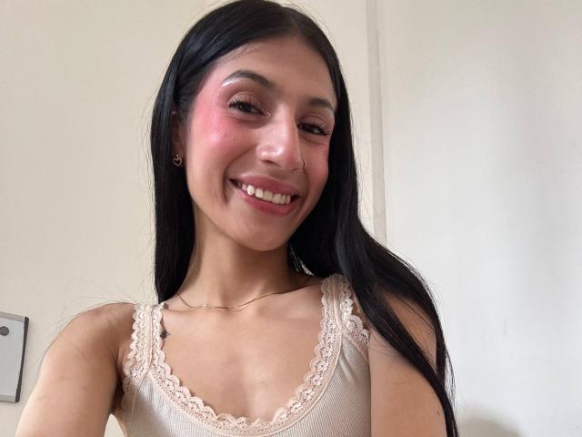 SophieBeaumont has long black hair, pink blush on her cheeks, and a cute smile. She's wearing a beige lace-trimmed top, showing her small tits. She's smiling at the camera.