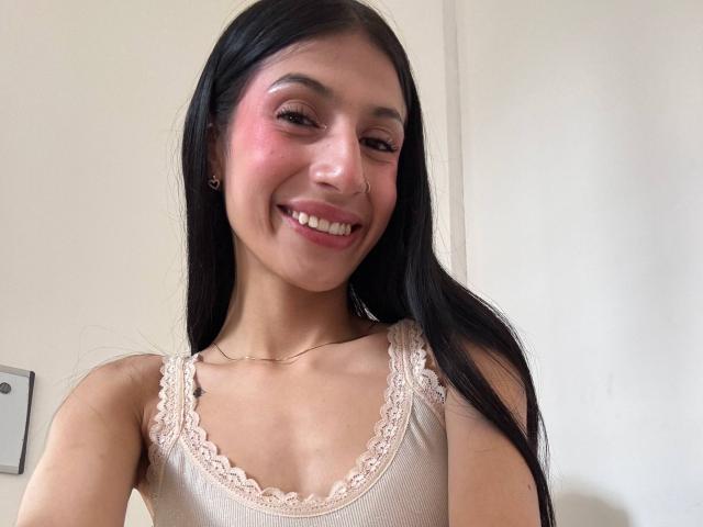 SophieBeaumont with long black hair, pink cheeks, smiling, wearing a lace-trimmed beige tank top, showing her small tits, and a gold necklace, in a selfie.