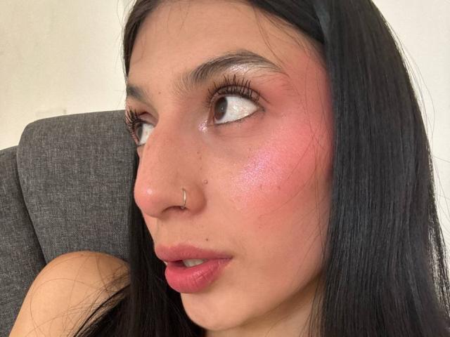SophieBeaumont has pinkish-red cheeks, shiny white eyelids, black hair, a nose ring, and pink lipstick. Her eyes are looking to the side, and she's sitting on a grey chair.