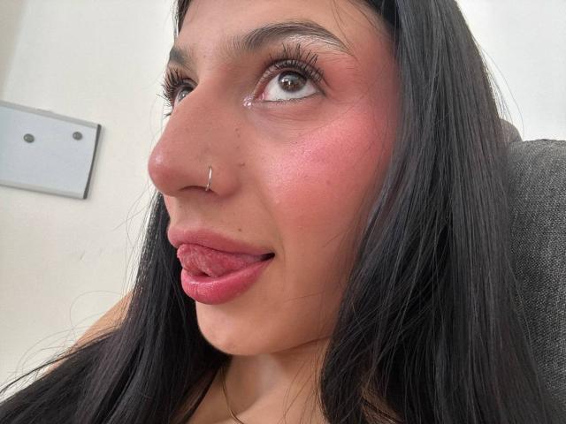 SophieBeaumont has long black hair, brown eyes, a silver nose ring, and is sticking her tongue out with pink lipstick. Her skin is slightly shiny and she's winking.