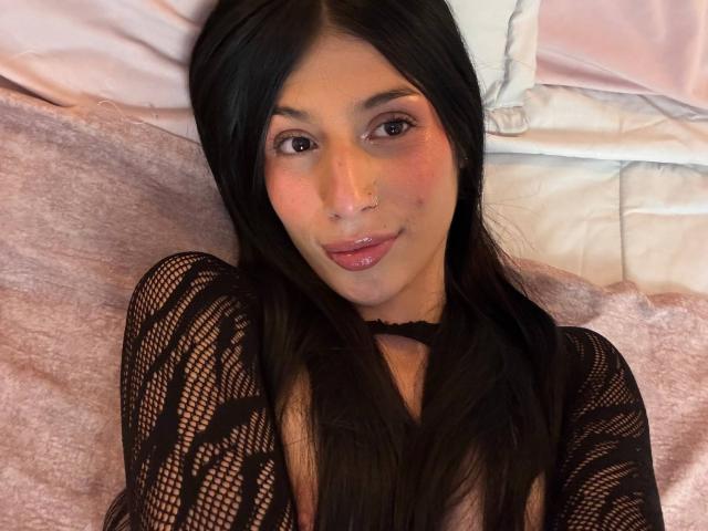 SophieBeaumont with long black hair, wearing a black lace top, has pinkish blush on her cheeks, a nose ring, and glossy pink lips, lying on a pink bed.
