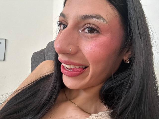 SophieBeaumont has long black hair, brown eyes, pink lipstick, nose ring, and a smile showing pink lipstick. She's wearing a lace top and gold earrings.