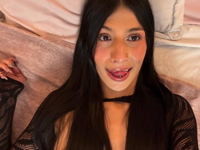 SophieBeaumont with long black hair, nose ring, and tongue piercing, sticking out her pink tongue, wearing a black lace top, lying on a bed.