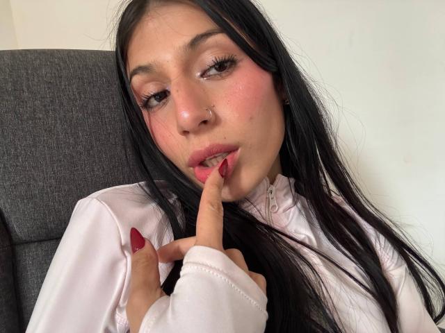 SophieBeaumont with long black hair, pink blush on her cheeks, nose ring, and red nails, touching her pink-lipped mouth with a finger, wearing a white shirt.