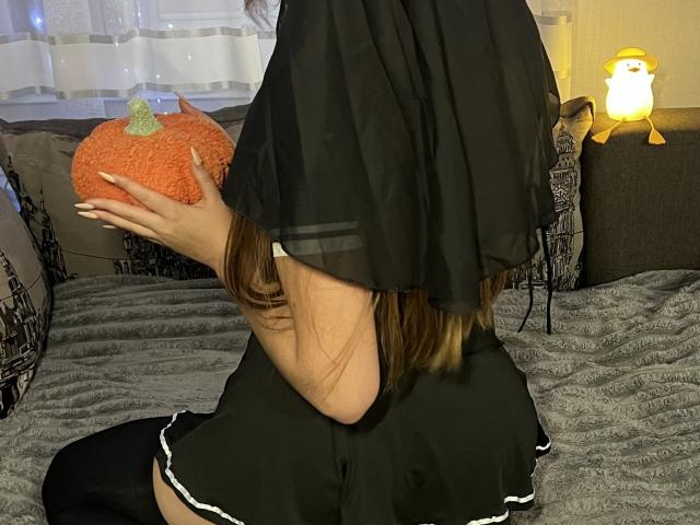 JoslynMensick with brown hair, black sheer dress, holding orange pumpkin, black skirt with white trim, black thigh-highs, sitting on gray bed, glowing penguin on right.